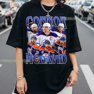 May include: Black t-shirt with a blue graphic of hockey player Connor McDavid and the text "Connor McDavid". The graphic includes images of McDavid and other players in their team uniforms.