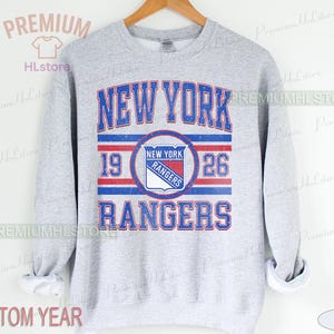 May include: A light grey sweatshirt with the text "NEW YORK RANGERS" in blue and red. The design includes the years "19" and "26" and a team logo. The sweatshirt is hanging on a wooden hanger.