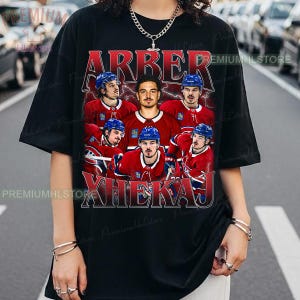 May include: Black t-shirt with a graphic of hockey players in red and blue. The text "ARBER XHEKAJ" is in a vintage style. The shirt has short sleeves and a crew neck. The image is a vintage style.