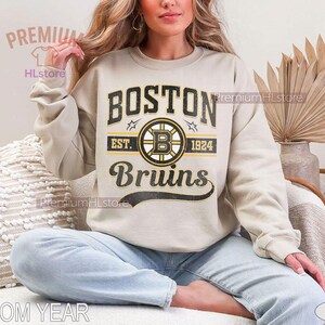 Puede incluir: Sudadera color arena con el texto "BOSTON Bruins" en negro y dorado, un logotipo de hockey y el a&ntilde;o 1924. La sudadera la lleva una persona sentada con las piernas cruzadas. Tambi&eacute;n se ven las palabras "CUSTOM YEAR" y "SAND".