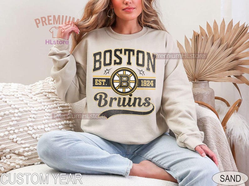 Puede incluir: Sudadera color arena con el texto "BOSTON Bruins" en negro y dorado, un logotipo de hockey y el a&ntilde;o 1924. La sudadera la lleva una persona sentada con las piernas cruzadas. Tambi&eacute;n se ven las palabras "CUSTOM YEAR" y "SAND".