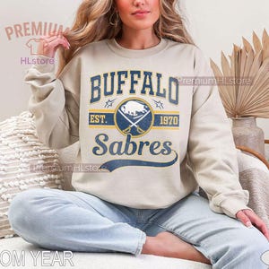 May include: A sand-colored sweatshirt featuring the Buffalo Sabres logo in navy blue and gold. The design includes the team name, a logo with crossed swords and a buffalo, and the text "EST. 1970". The words "CUSTOM YEAR" are visible at the bottom.