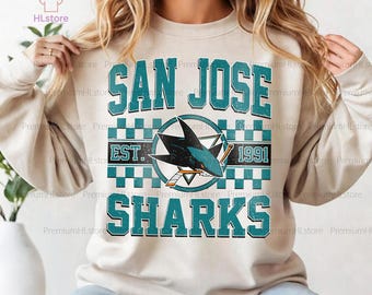 May include: A sand-coloured sweatshirt with the teal text "SAN JOSE SHARKS". The design includes a shark logo with a hockey stick and the year "1991". The sweatshirt has a crew neck and long sleeves.