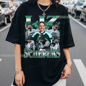 Könnte beinhalten: Schwarzes T-Shirt mit einem grün-weißen Grafikdesign eines Hockeyteams, einschließlich des Namens "LIZ SCHEPERS". Das Design zeigt ein zentrales Porträt und andere Spieler in Hockey-Uniformen.