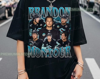 May include: Black t-shirt featuring a graphic of hockey player Brandon Montour. The design includes the player's name and images of him in his team uniform. The text is in a teal and white color scheme.