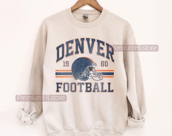 May include: A beige sweatshirt featuring the text "DENVER FOOTBALL" in blue. A blue football helmet is centered below the word "DENVER". The year "1960" is above the helmet. Two orange stripes are below the helmet.