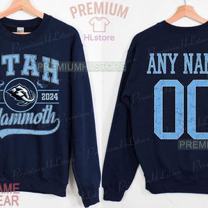 K&ouml;nnte beinhalten: Marineblaues Sweatshirt mit "UTAH" in Hellblau, einem Mammoth-Logo und "EST. 2024". Auf der R&uuml;ckseite stehen "ANY NAME" und die Nummer "00" in Hellblau. Ein individueller Name und eine Nummer k&ouml;nnen hinzugef&uuml;gt werden.
