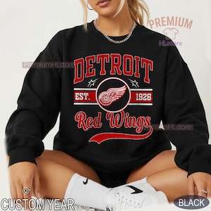 Puede incluir: Sudadera negra con la palabra "DETROIT" en letras rojas sobre "Red Wings". El dise&ntilde;o incluye un emblema circular con una rueda alada y el texto "EST. 1926". La sudadera tiene cuello redondo y mangas largas.