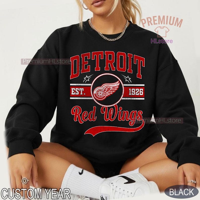 Puede incluir: Sudadera negra con la palabra "DETROIT" en letras rojas sobre "Red Wings". El dise&ntilde;o incluye un emblema circular con una rueda alada y el texto "EST. 1926". La sudadera tiene cuello redondo y mangas largas.