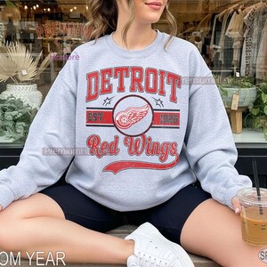 Puede incluir: Sudadera gris claro con la inscripci&oacute;n "DETROIT Red Wings" en rojo y un logotipo de rueda alada. La sudadera tambi&eacute;n muestra "EST 1926". Los textos "CUSTOM YEAR" y "SPORT GREY" tambi&eacute;n son visibles.