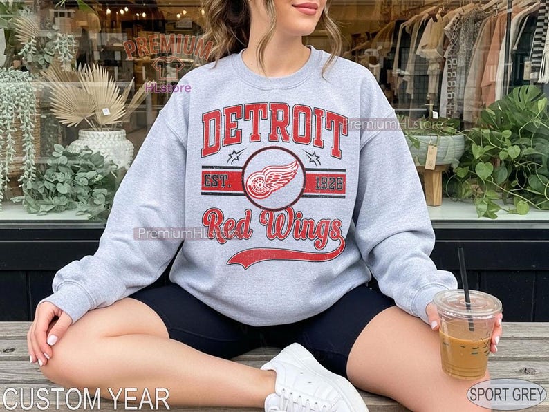 Puede incluir: Sudadera gris claro con la inscripci&oacute;n "DETROIT Red Wings" en rojo y un logotipo de rueda alada. La sudadera tambi&eacute;n muestra "EST 1926". Los textos "CUSTOM YEAR" y "SPORT GREY" tambi&eacute;n son visibles.