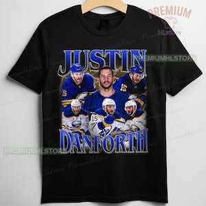 May include: Black t-shirt featuring a collage of hockey players in blue and white uniforms. The shirt displays the name "JUSTIN DANFORTH" in a blue, vintage-style font. The design includes a central portrait of a man.