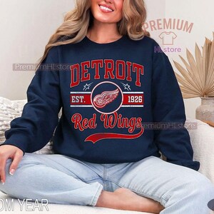 Puede incluir: Sudadera azul marino con el texto "DETROIT Red Wings" en rojo y blanco, y un logotipo de rueda alada. La sudadera tambi&eacute;n incluye "EST. 1926". Una prenda informal para los aficionados al hockey sobre hielo.