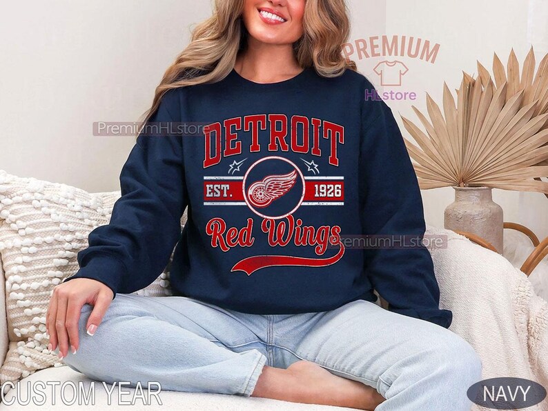 Puede incluir: Sudadera azul marino con el texto "DETROIT Red Wings" en rojo y blanco, y un logotipo de rueda alada. La sudadera tambi&eacute;n incluye "EST. 1926". Una prenda informal para los aficionados al hockey sobre hielo.