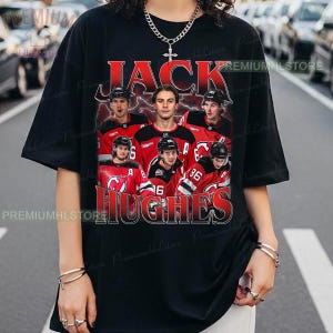 May include: Black t-shirt featuring a graphic of a hockey player with the name "JACK HUGHES" in red and white lettering. The design includes images of the player and other team members in red jerseys.