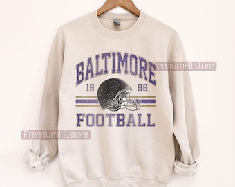 Puede incluir: Sudadera beige con la inscripción "BALTIMORE FOOTBALL" en morado, un casco de fútbol americano y el año "1996". La sudadera está colgada de una percha de madera.