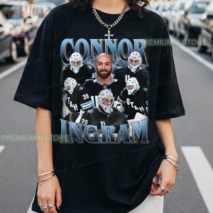 May include: A black short-sleeve t-shirt featuring a graphic print of a hockey player named Connor Ingram. The design includes a central portrait of the player surrounded by four images of him in a black and white hockey uniform and helmet.