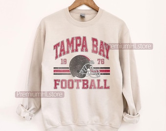 Puede incluir: Sudadera beige claro con un diseño gráfico desgastado. El gráfico dice "TAMPA BAY FOOTBALL" en rojo, con un casco de fútbol americano y los números "19" y "76". La sudadera está colgada de una percha de madera.