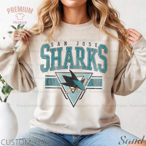 May include: Beige sweatshirt featuring the San Jose Sharks logo in teal and black. The design includes the team name, a shark graphic, and the years "EST. 1991". The sweatshirt has long sleeves and a crew neck.