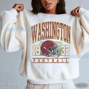 May include: Cream-coloured sweatshirt with a maroon and gold football helmet graphic. The text "WASHINGTON 1932 FOOTBALL" is printed above and below the helmet. The sweatshirt has long sleeves and a crew neck.