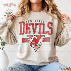 May include: A sand-colored sweatshirt featuring the text "NEW JERSEY DEVILS" in a distressed red font. Below the text is a logo with a red devil and the text "EST. 1974". The sweatshirt has long sleeves and a crew neck.