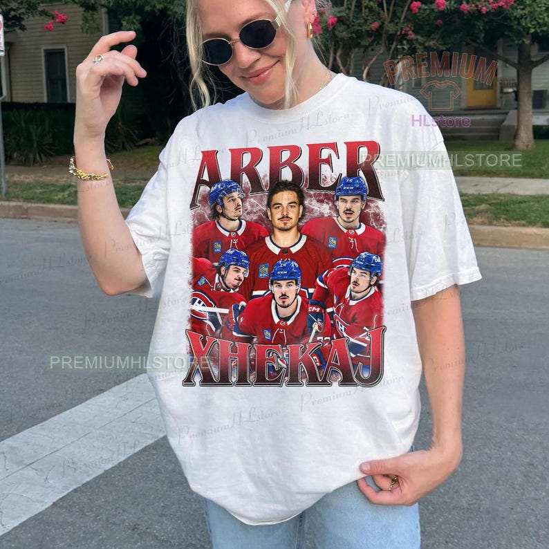 May include: White t-shirt featuring a graphic of a hockey team in red and blue uniforms. The word "ARBER" is printed above the players, and "XHEKAJ" is printed below. The shirt has short sleeves.
