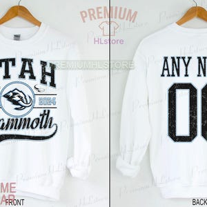 K&ouml;nnte beinhalten: Wei&szlig;es Sweatshirt mit "UTAH Mammoth" Text und Logo auf der Vorderseite, mit "EST. 2004" und einer Mammut-Grafik. Auf der R&uuml;ckseite steht "ANY NAME" &uuml;ber der Nummer "00". Das Sweatshirt wird auf einem Holzb&uuml;gel pr&auml;sentiert.