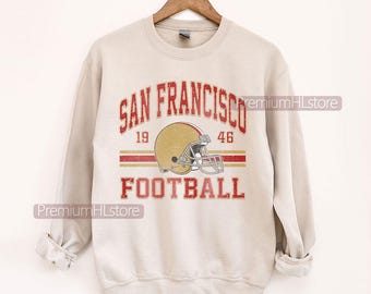 May include: A light beige sweatshirt with a distressed graphic. The graphic features the words "SAN FRANCISCO FOOTBALL" in red, with a gold football helmet and the numbers "19" and "46".