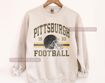 May include: A light beige sweatshirt featuring a distressed graphic of a football helmet and the words "PITTSBURGH FOOTBALL" in black and gold. The sweatshirt has a crew neck and long sleeves. The text "1933" is also included in the design.