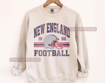 以下が含まれることがあります： ベージュ色のスウェットシャツ。青と赤の文字で「NEW ENGLAND FOOTBALL」と書かれています。フットボールのヘルメットのグラフィックがテキストの下に配置されています。スウェットシャツは木製のハンガーに掛けられています。