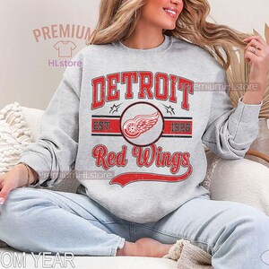 Puede incluir: Una sudadera gris claro con la inscripci&oacute;n "DETROIT Red Wings" en rojo, y un logotipo de rueda alada. La sudadera tambi&eacute;n tiene el texto "EST 1926". El dise&ntilde;o es de inspiraci&oacute;n vintage.