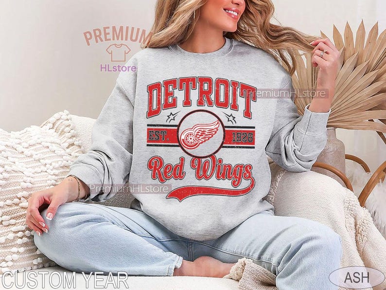 Puede incluir: Una sudadera gris claro con la inscripci&oacute;n "DETROIT Red Wings" en rojo, y un logotipo de rueda alada. La sudadera tambi&eacute;n tiene el texto "EST 1926". El dise&ntilde;o es de inspiraci&oacute;n vintage.