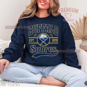 May include: Navy blue sweatshirt featuring the text "BUFFALO Sabres" in gold and white lettering. The design includes a logo with crossed hockey sticks and a buffalo. The year "1970" is also displayed.