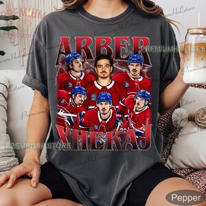 May include: Dark gray t-shirt featuring a graphic of a hockey player in a red and blue uniform, surrounded by other players. The name "ARBER XHEKAJ" is printed in red across the top and bottom of the graphic. The shirt has a vintage, distressed look.