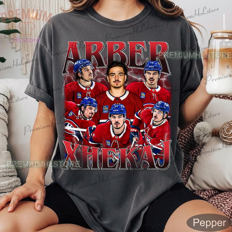 May include: Dark gray t-shirt featuring a graphic of a hockey player in a red and blue uniform, surrounded by other players. The name "ARBER XHEKAJ" is printed in red across the top and bottom of the graphic. The shirt has a vintage, distressed look.