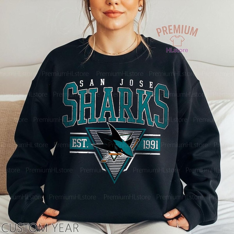 May include: Black sweatshirt featuring the text "SAN JOSE SHARKS" in teal and white. Below the text is a logo of a shark with hockey sticks and the year "1991". The sweatshirt has a crew neck and long sleeves.