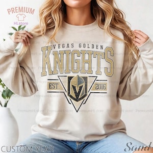 Può includere: Una felpa beige con la scritta "VEGAS GOLDEN KNIGHTS" in oro e nero. Sotto, uno stemma con la scritta "EST. 2016". La felpa ha maniche lunghe e girocollo.