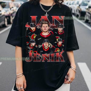 Puede incluir: Camiseta negra con un gráfico rojo de un jugador de hockey y el texto "JAN JENIK". El diseño incluye múltiples imágenes del jugador con su uniforme. La camiseta tiene cuello redondo y mangas cortas.