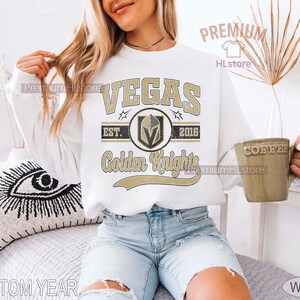Puede incluir: Sudadera blanca con el texto "VEGAS Golden Knights" en dorado y negro. El dise&ntilde;o incluye un logotipo del equipo y el a&ntilde;o 2016. La sudadera es de estilo casual.