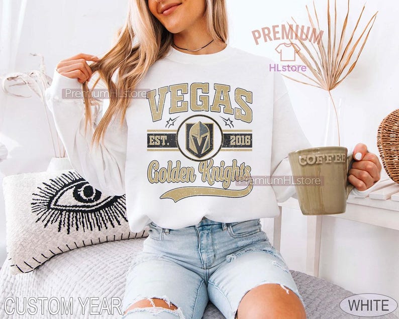 Puede incluir: Sudadera blanca con el texto "VEGAS Golden Knights" en dorado y negro. El dise&ntilde;o incluye un logotipo del equipo y el a&ntilde;o 2016. La sudadera es de estilo casual.