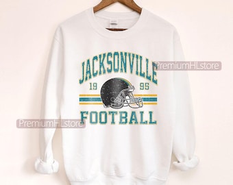 May include: White sweatshirt with a distressed graphic of a football helmet and the text "JACKSONVILLE FOOTBALL 1995". The design includes teal and gold accents. The sweatshirt is displayed on a wooden hanger.