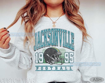 May include: Pale grey sweatshirt featuring a teal and white graphic reading "JACKSONVILLE 1995 FOOTBALL". The design includes a football helmet. The words "CUSTOM YEAR" are at the bottom left.