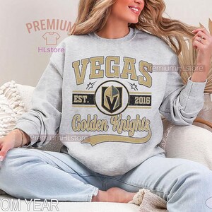 Puede incluir: Sudadera gris claro con la palabra "VEGAS" en letras doradas. Debajo, un emblema circular con un casco de caballero y las palabras "Golden Knights". La sudadera tambi&eacute;n tiene el texto "EST. 2016".