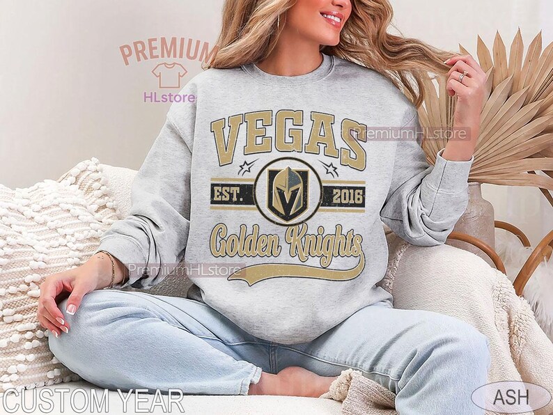 Puede incluir: Sudadera gris claro con la palabra "VEGAS" en letras doradas. Debajo, un emblema circular con un casco de caballero y las palabras "Golden Knights". La sudadera tambi&eacute;n tiene el texto "EST. 2016".