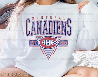 Puede incluir: Sudadera blanca de manga larga con el texto "MONTREAL CANADIENS" en azul y rojo, con un logotipo y "EST. 1909". El diseño tiene un aspecto desgastado y vintage. Es una prenda deportiva informal.