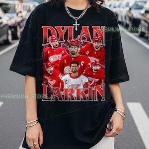 May include: Black t-shirt featuring a graphic of Dylan Larkin and other hockey players in red jerseys. The text "DYLAN LARKIN" is displayed in red, with a distressed effect. The shirt has a crew neck and short sleeves.