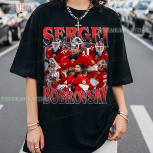 May include: Black t-shirt featuring a graphic of a hockey team in red jerseys. The text "SERGEI BOBROVSKY" is printed in red and white. The shirt has a crew neck and short sleeves.