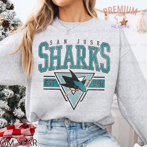 May include: A light gray sweatshirt featuring the text "SAN JOSE SHARKS" in teal block letters. Below the text is a teal and black shark logo with the text "EST 1991". The words "CUSTOM YEAR" are at the bottom.
