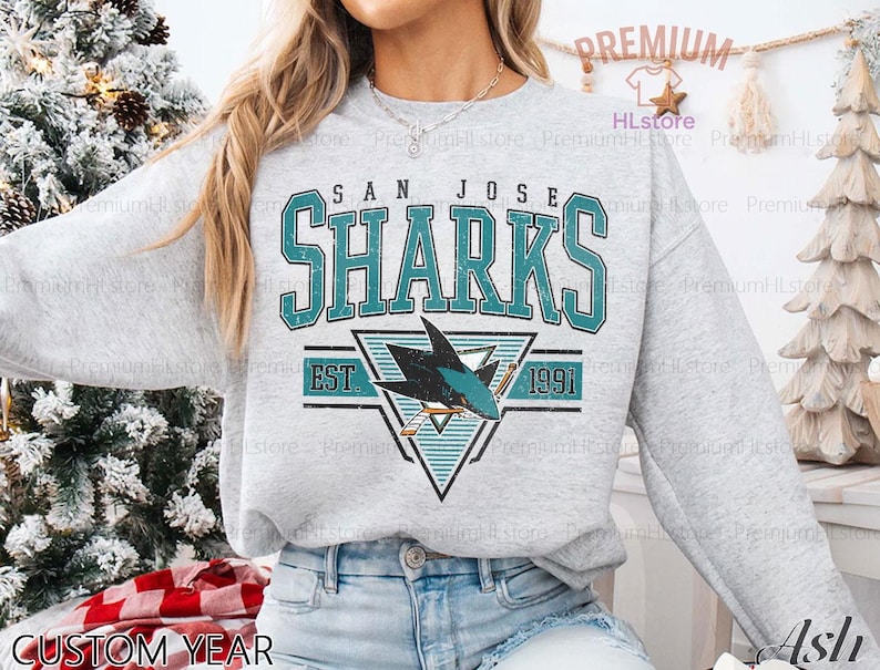 May include: A light gray sweatshirt featuring the text "SAN JOSE SHARKS" in teal block letters. Below the text is a teal and black shark logo with the text "EST 1991". The words "CUSTOM YEAR" are at the bottom.