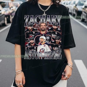 May include: Black t-shirt featuring a graphic of hockey players with the names "ZACHARY" and "ASTON REESE" in a vintage style. The design includes images of players in their team uniforms. The shirt has a crew neck and short sleeves.